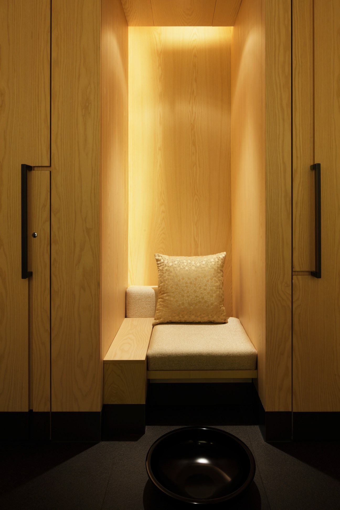 Minimalist spa therapy room with wooden walls, illuminated niche, cushioned bench seating and black bowl.