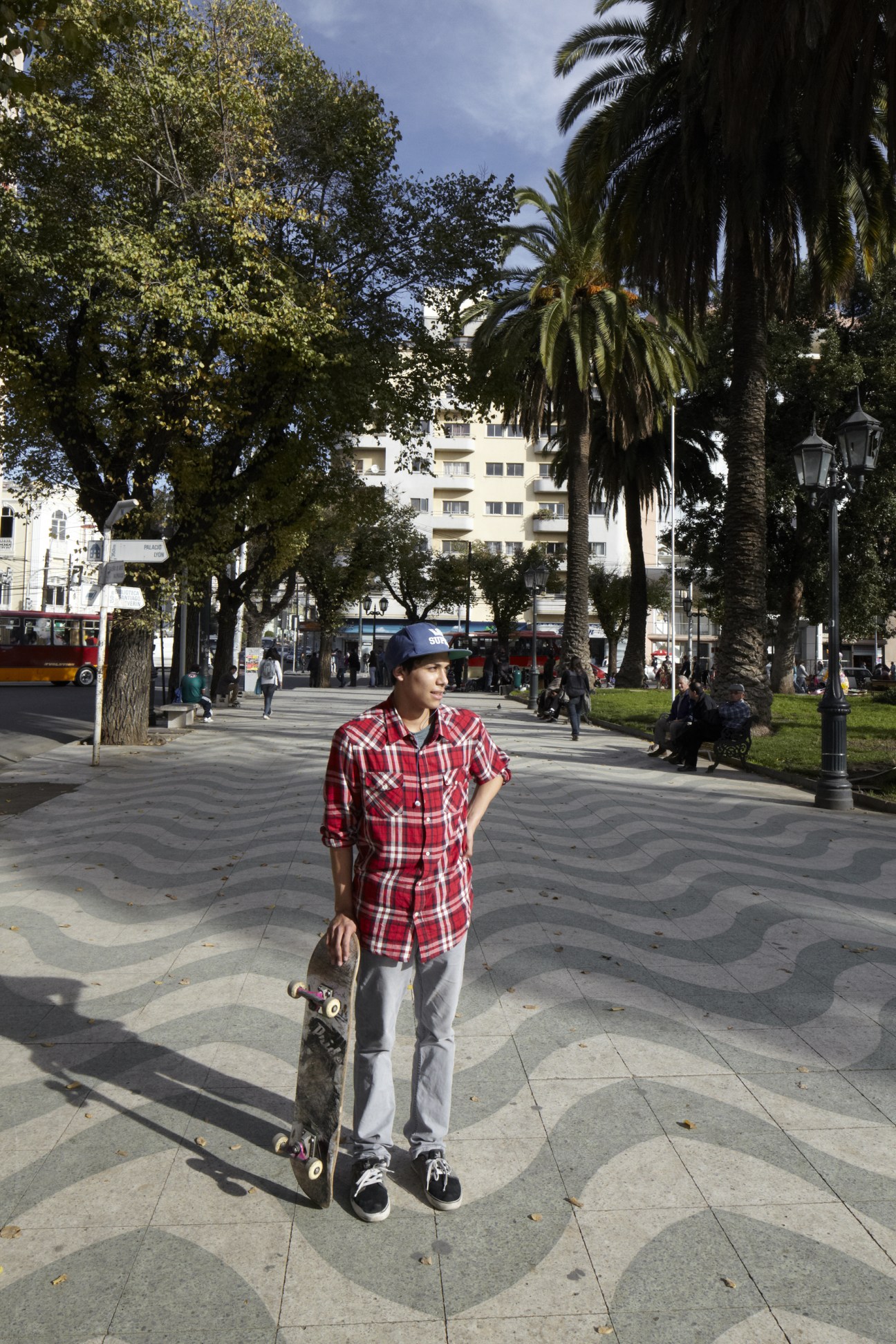 Young skateboarder in red plaid shirt standing at Plaza Victoria in Valparaíso with palm trees and white buildings in background.