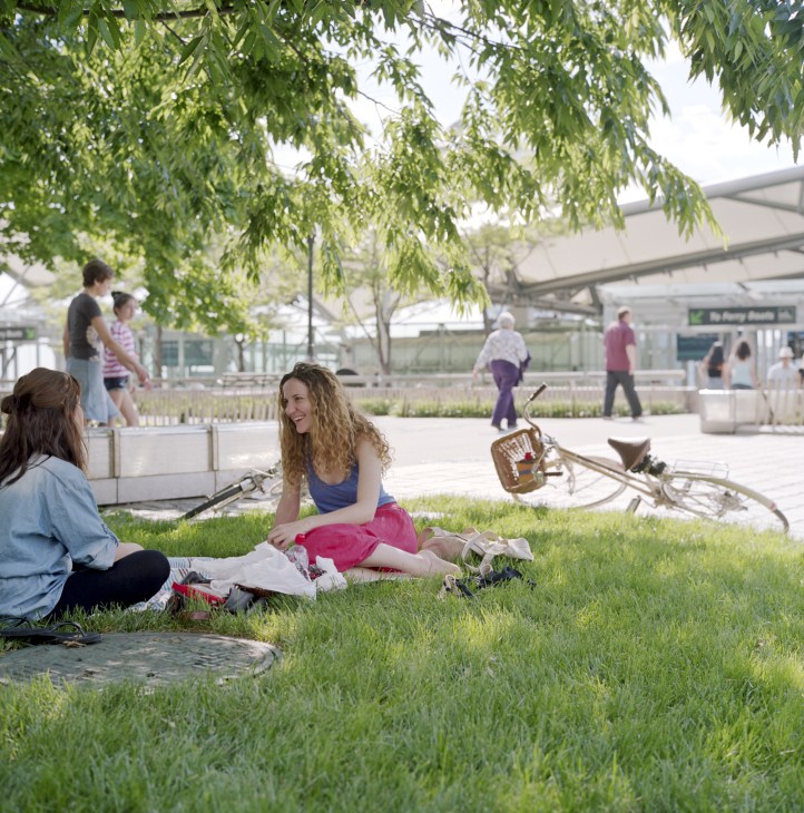 Two women sit on grass in a public park with people and a bicycle visible in the background.