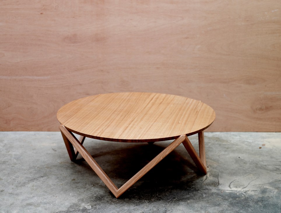 Circular wooden coffee table with light wood top and geometric dark wood base on concrete floor.