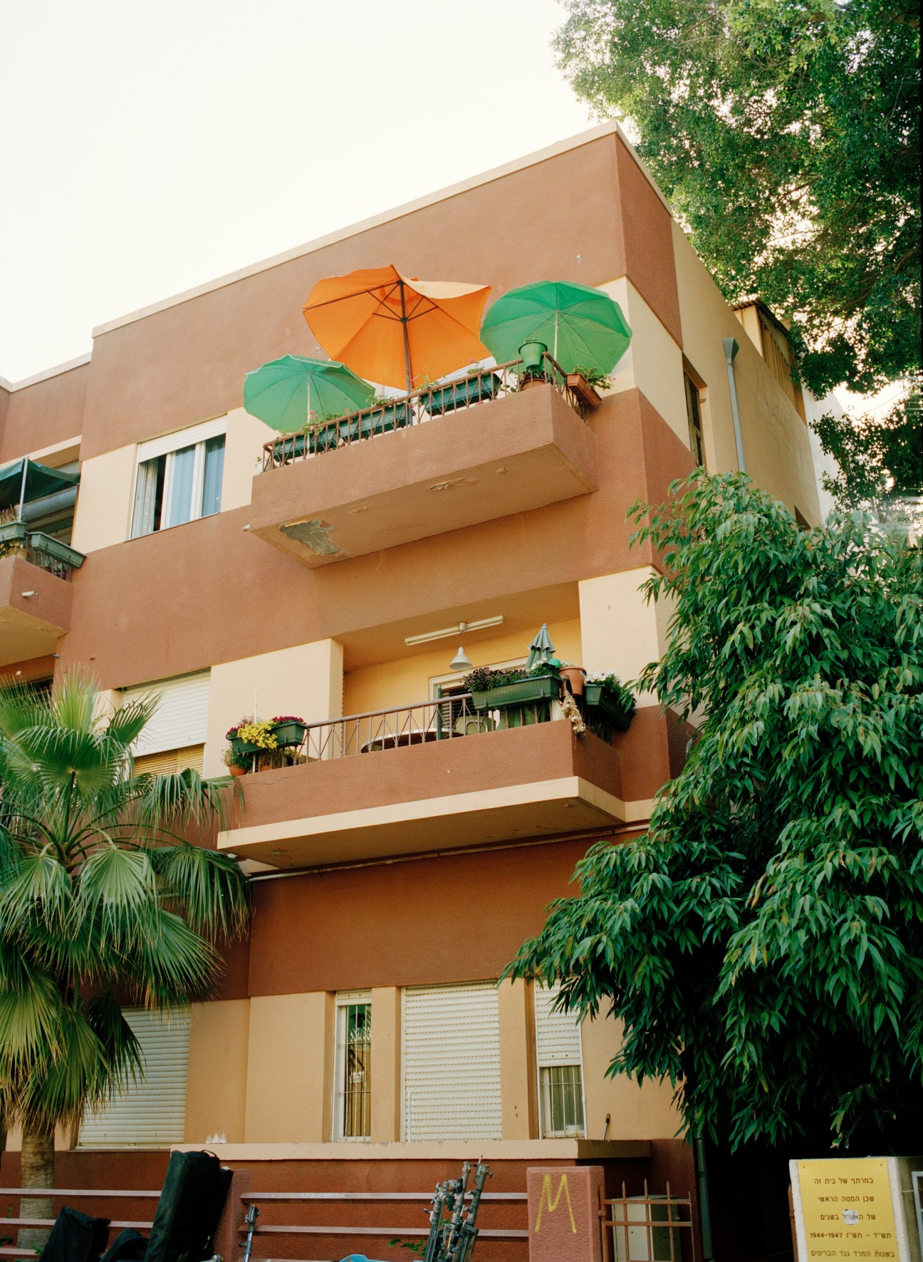 Terracotta-coloured Bauhaus building with multiple balconies featuring colourful umbrellas and surrounding green vegetation.