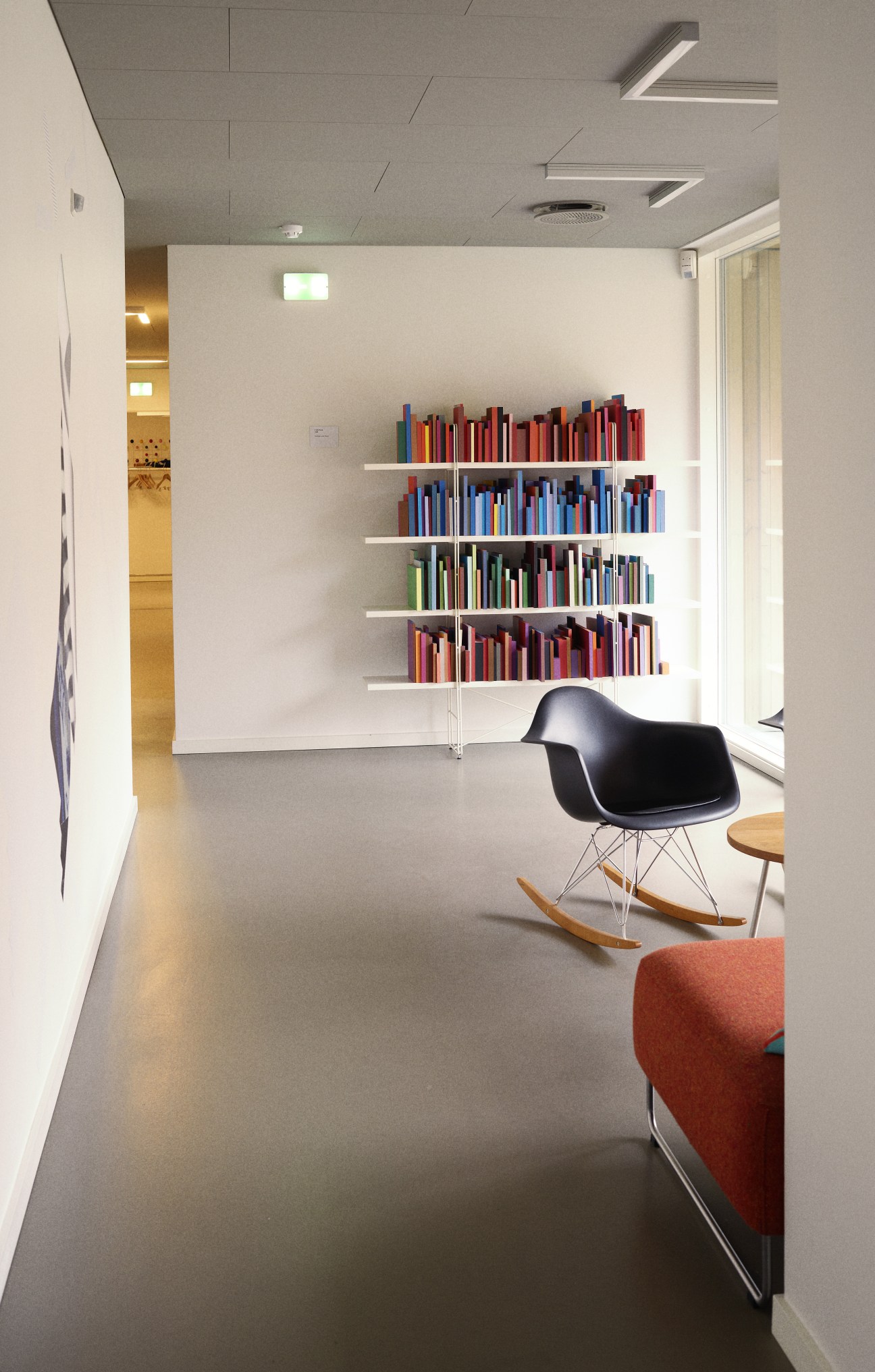 A quiet hallway nook with an Eames rocking chair, colorful books on wall shelves, and natural light from a window.