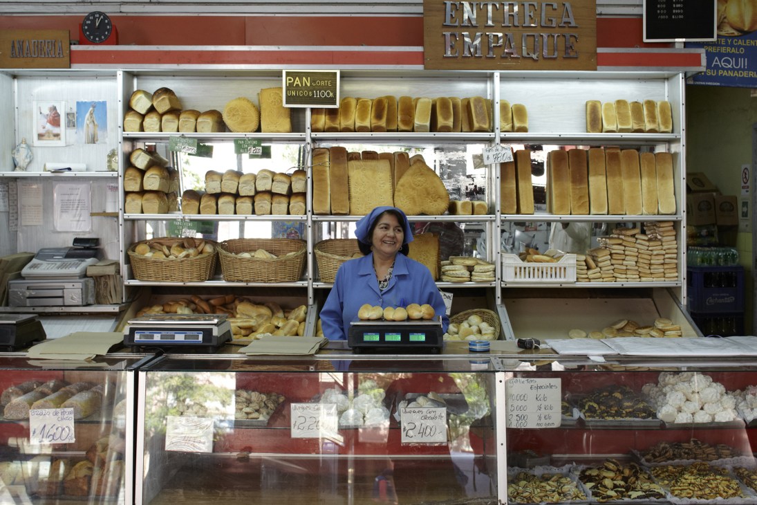 Baker in blue uniform stands at counter of traditional bakery with shelves of loaves and baked goods behind her.