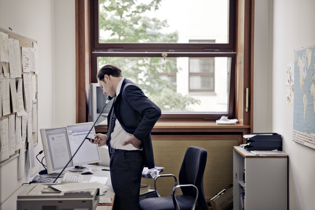 A man in a dark suit speaks on a telephone at his desk in a modest office with large windows.
