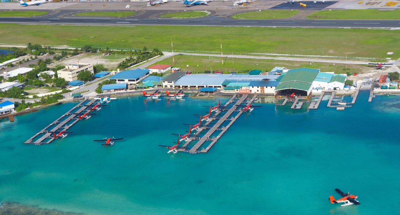 Aerial view of Maldivian Air Taxi seaplane base with red and white aircraft docked at wooden piers in turquoise water.