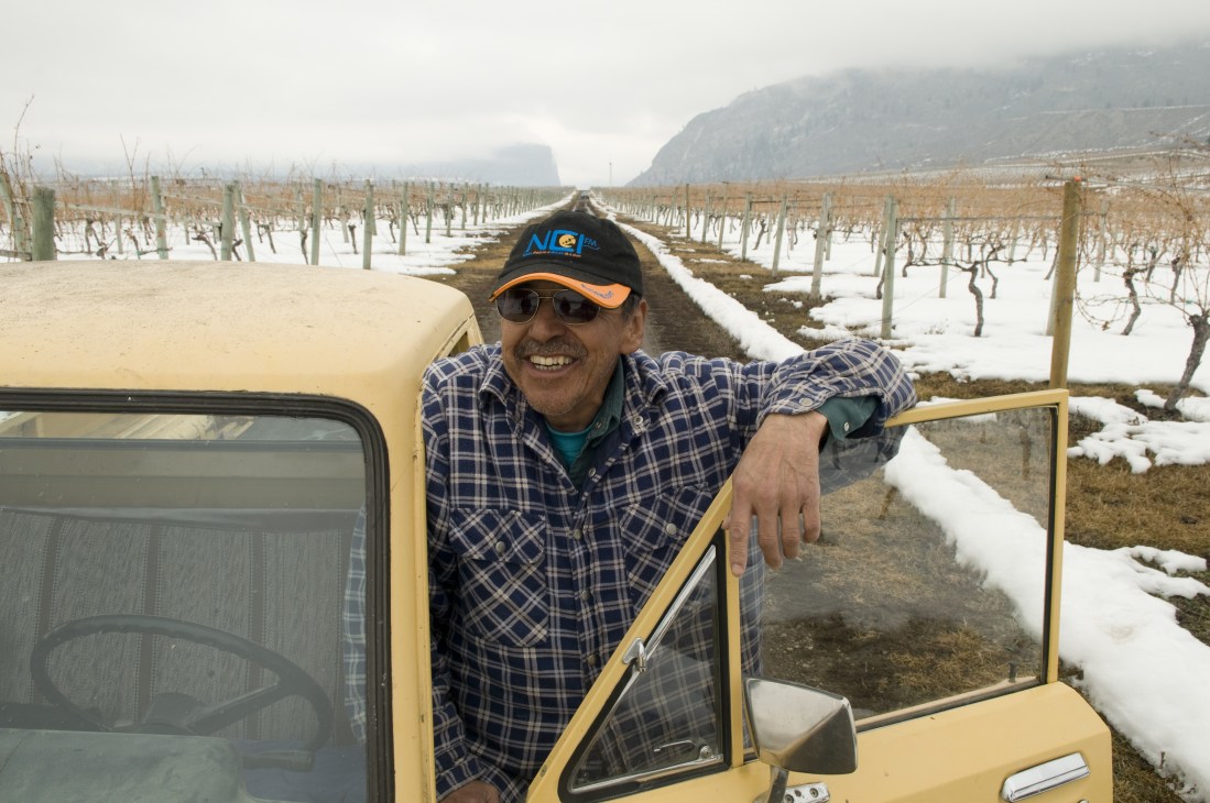 Vine pruner Steve Louie Lezard smiling from a yellow vehicle overlooking snow-covered Nk'Mip Vineyard rows.