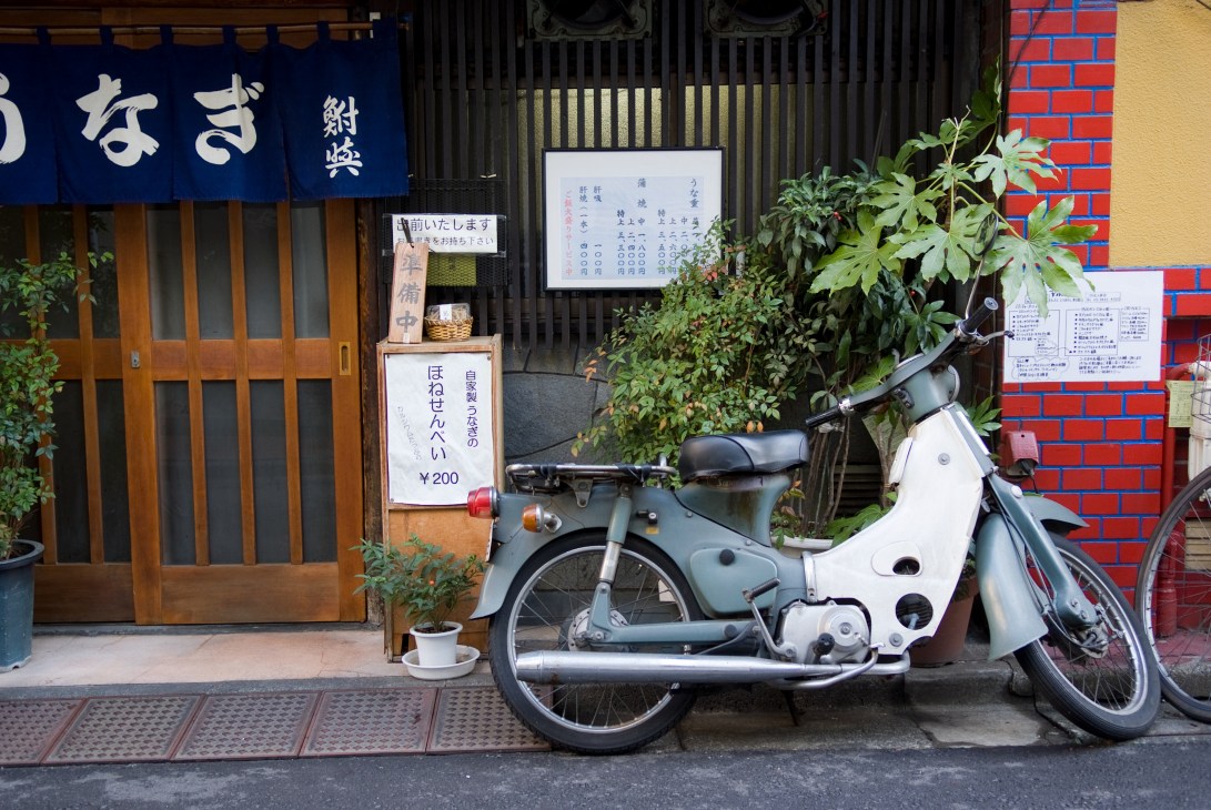 White and grey Honda Super Cub scooter parked outside a traditional Japanese restaurant with wooden storefront.