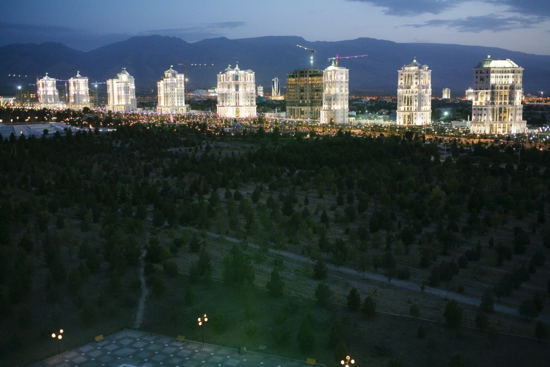 Illuminated marble apartment buildings at dusk in Ashgabat with mountains in the background.
