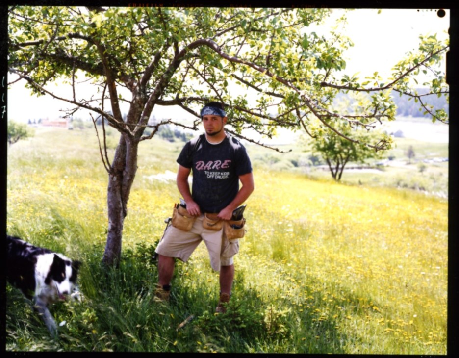 Man in vintage DARE t-shirt and tool belt stands under tree in sunny field landscape.