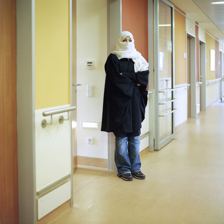 Woman wearing white headscarf and black coat stands in hospital corridor with yellow and orange walls.