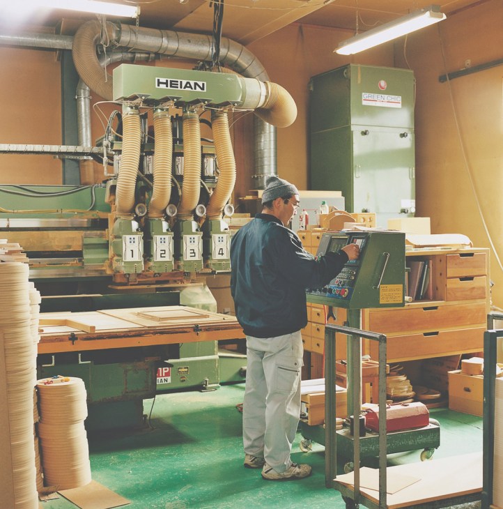 Woodworker operating a large Heian carving machine in a workshop, with wood planks and dust collection system visible.