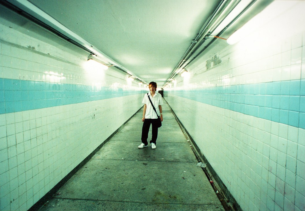 A young person in a white shirt stands alone in a long, turquoise-tiled underground tunnel with fluorescent lighting.