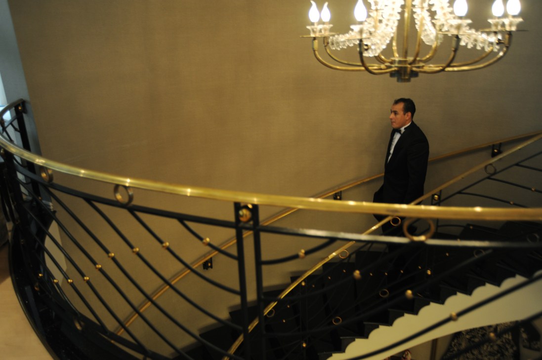 A man in a dark suit stands on a curved staircase with gold-trimmed railings in an elegant interior space.