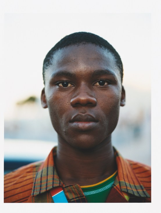 Portrait of a young man from Mozambique wearing an orange and green plaid shirt against a blurred background.