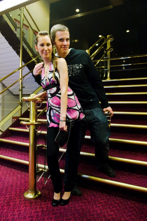 Two people smiling on an illuminated staircase with brass railings inside a ship.