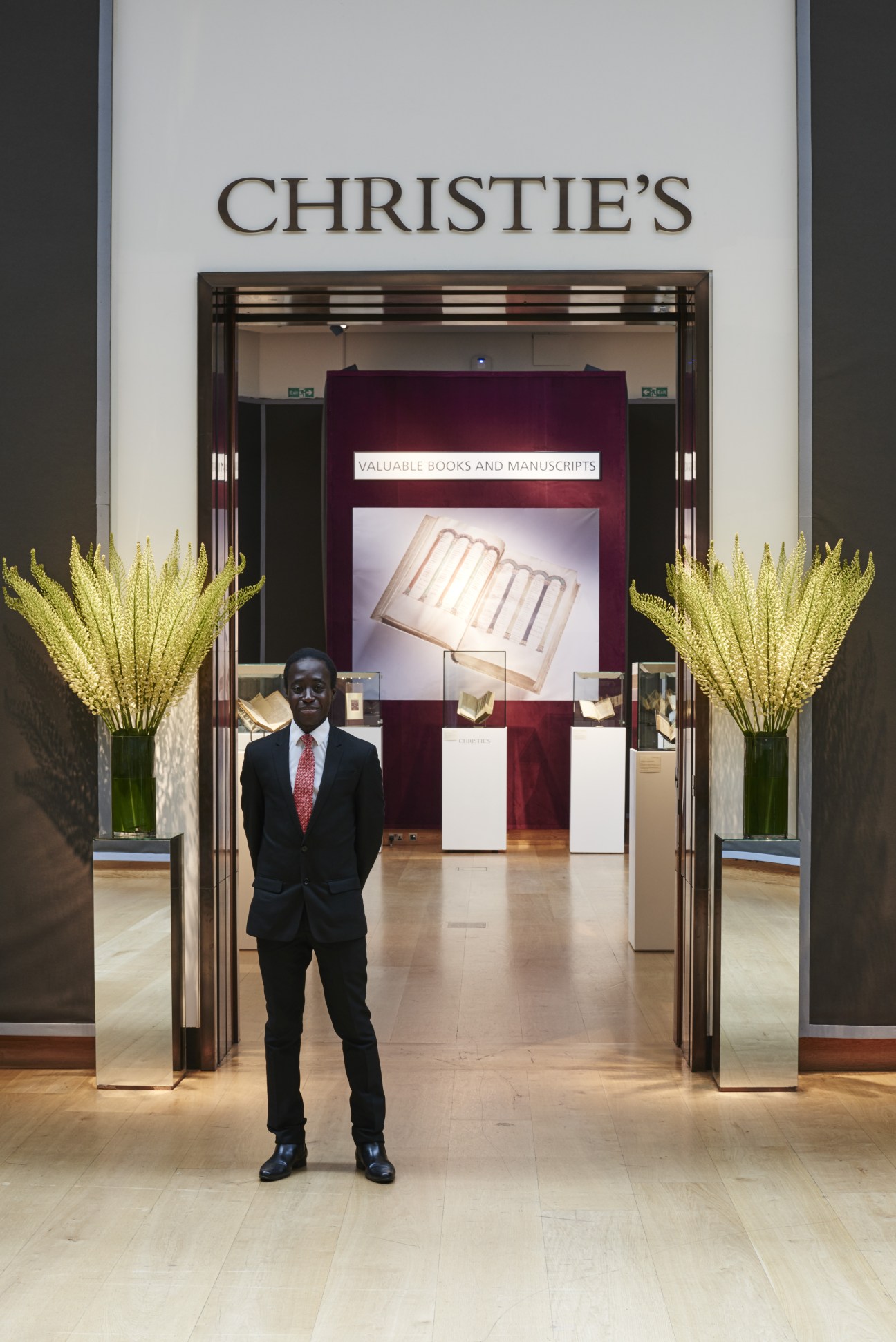 Man in dark suit standing in Christie's auction gallery with valuable books display.