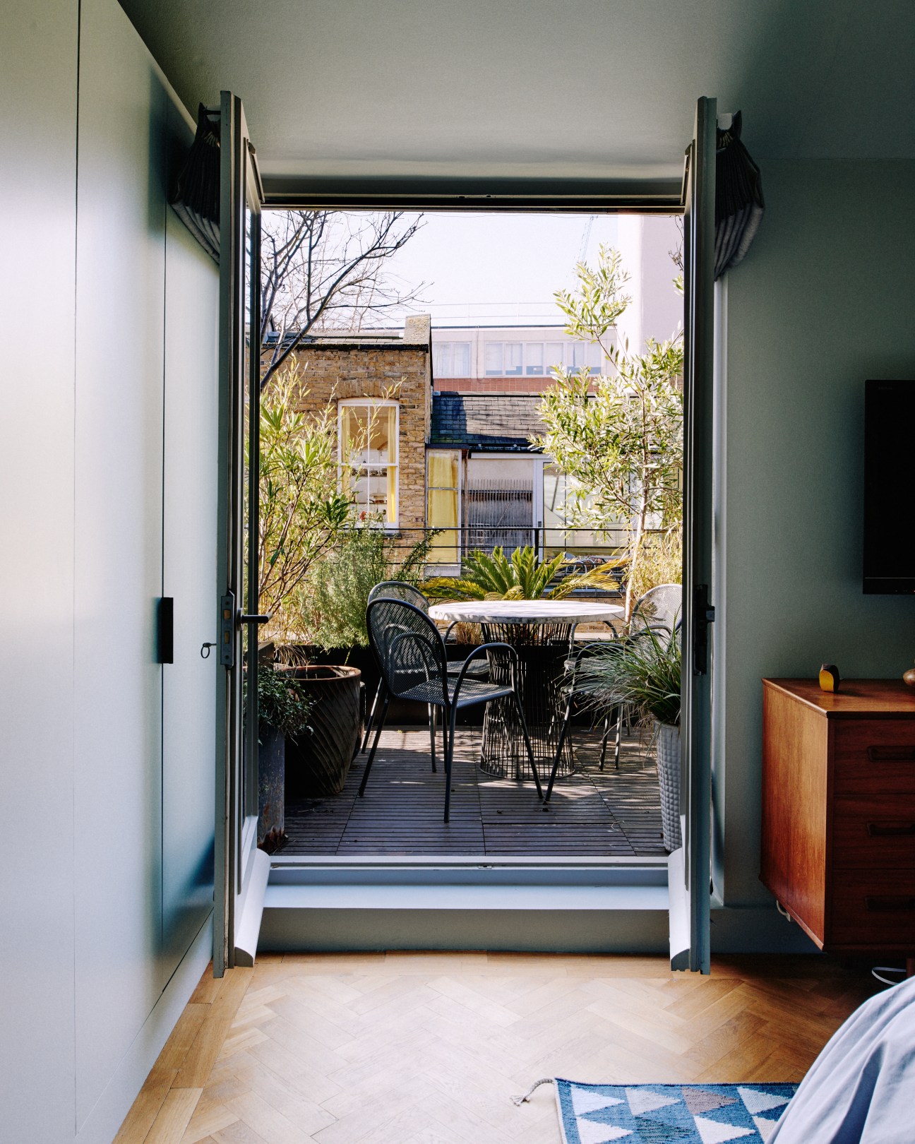 View from a bedroom through open glass doors to a small roof terrace with black chairs and potted plants.