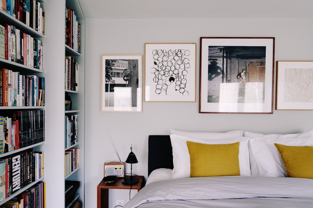 Bedroom with framed artwork on white walls, bookshelves, bed with yellow pillows, and minimal furnishings.
