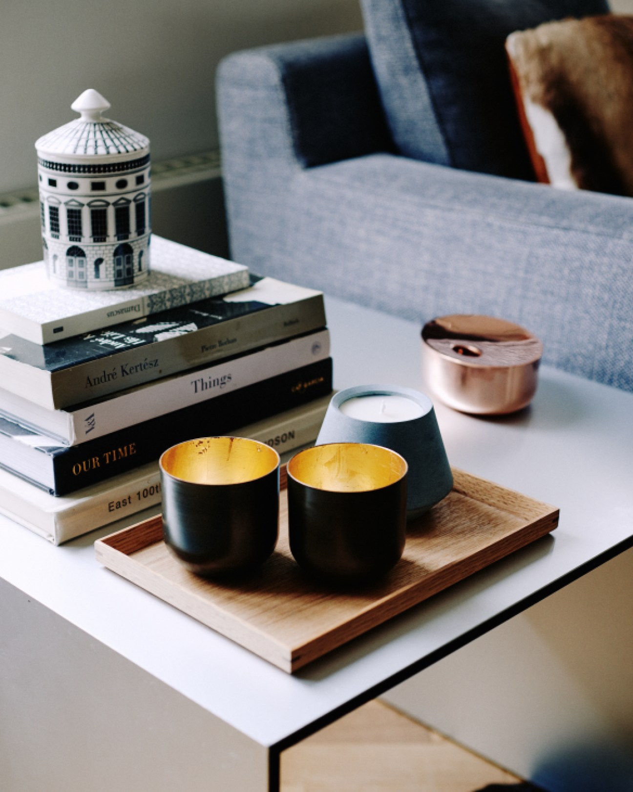 Black tea lights with gold interiors arranged on a wooden tray next to books and a blue chair.