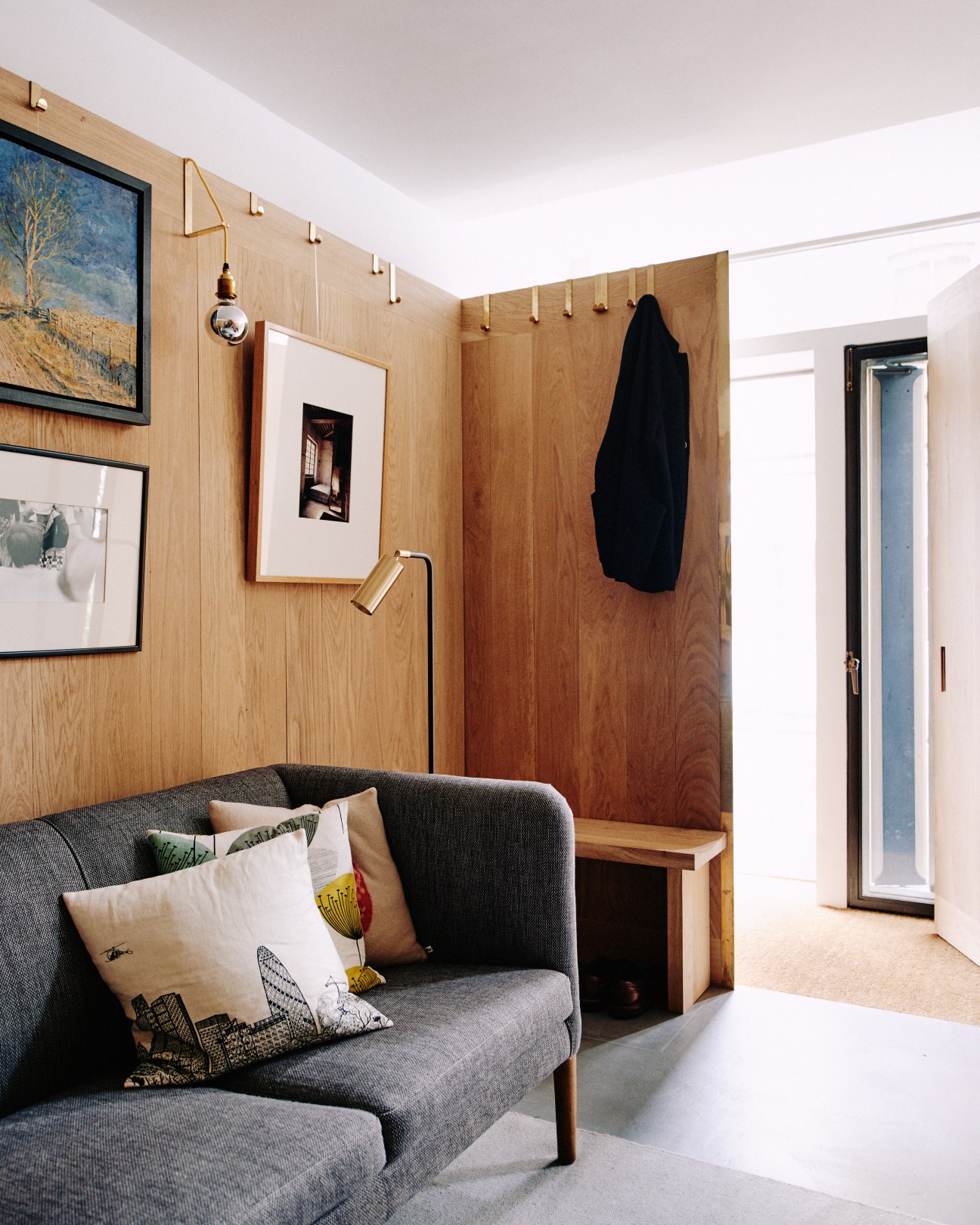 Ground floor study with oak-paneled walls, gray sofa with cushions, brass pendant lights, framed artwork, and wooden bench near entrance.