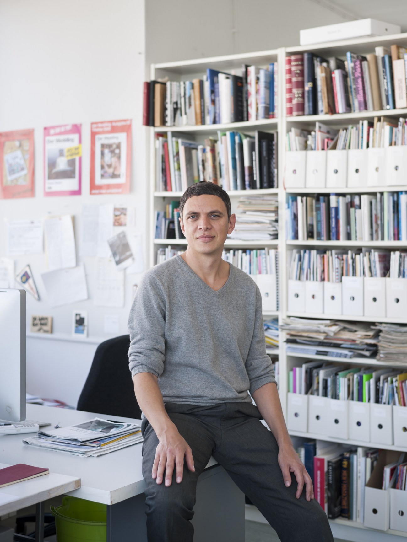 Man in grey sweater sitting at desk in front of white shelving unit filled with books and magazines.