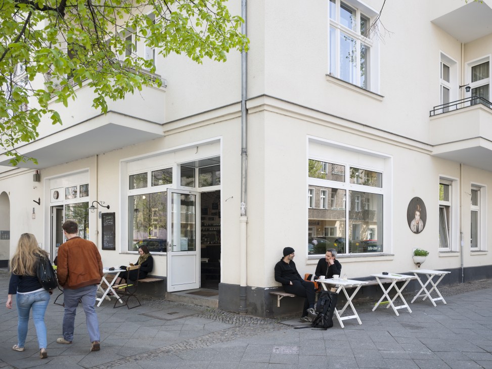 Exterior of TassenKuchen café with white seating and people outside the storefront on Malplaquetstrasse.