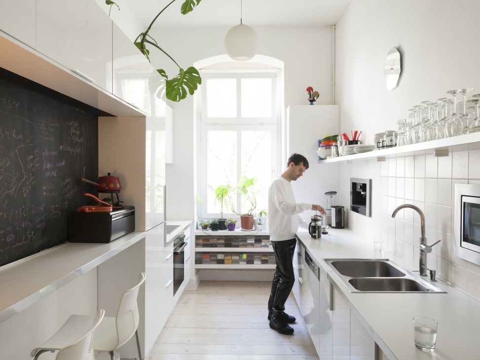 Greg Blunt standing in a bright, minimalist kitchen with white cabinetry, black chalkboard wall, and plants.