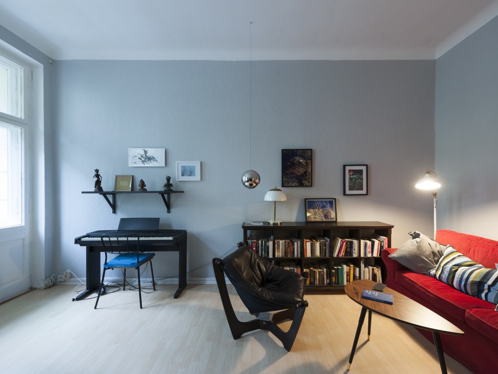 A bright modern apartment interior with blue-gray walls, a red sofa, black keyboard, bookshelf and curated artwork.