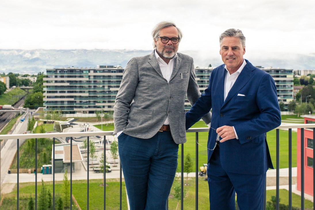 Two men stand on a balcony overlooking a green park with modern buildings and mountains in the background.