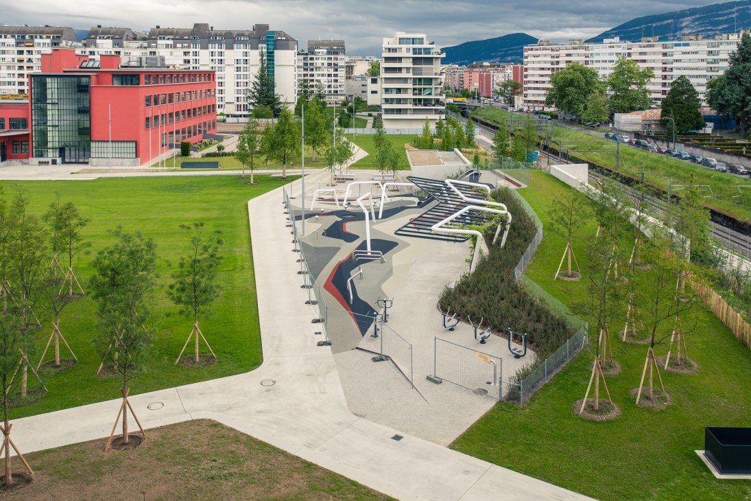 Aerial view of a modern Geneva park with geometric pathways, young trees, green lawns, and a red building nearby with mountains in the background.