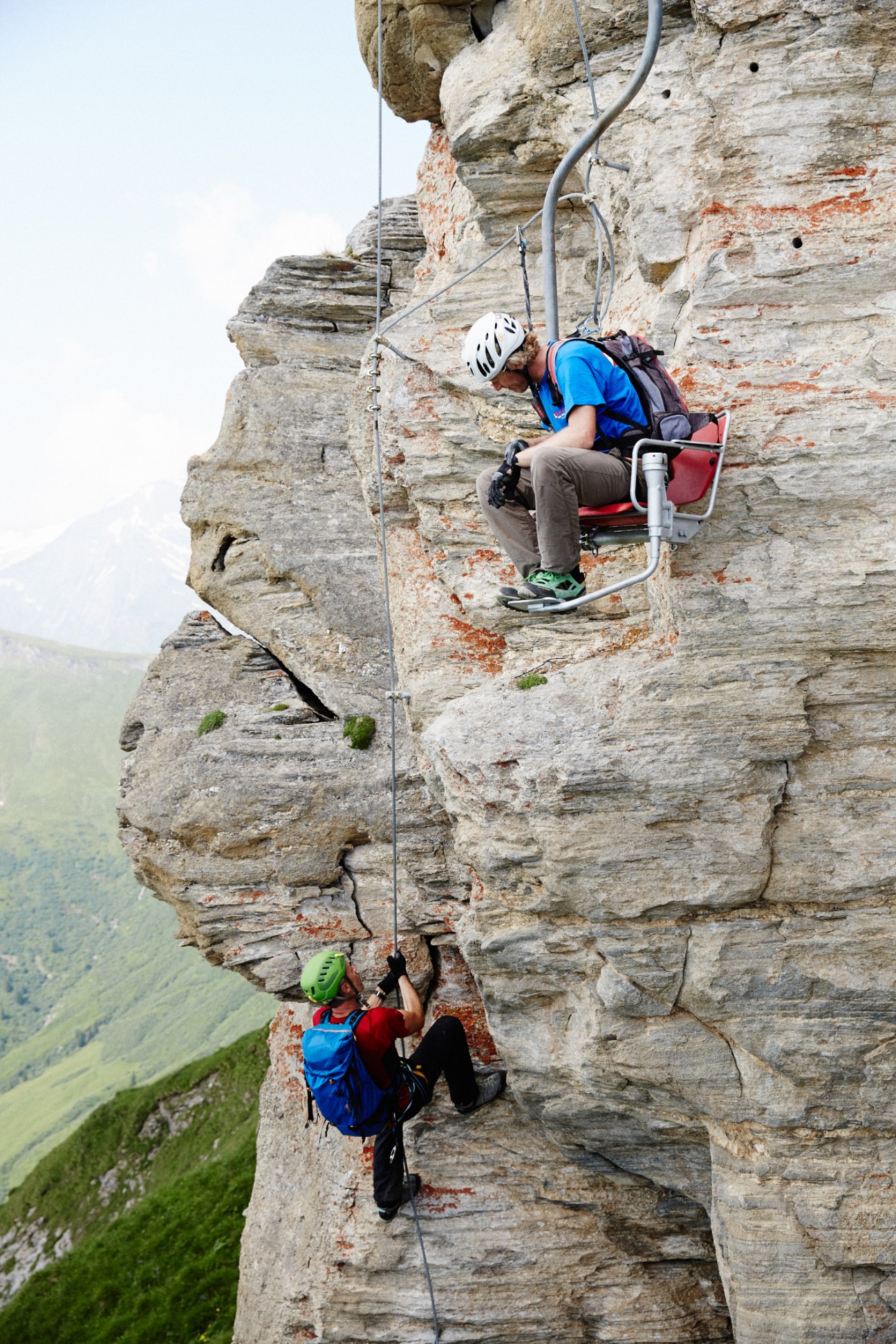Two climbers scale a rocky cliff face with safety equipment in an Alpine landscape.
