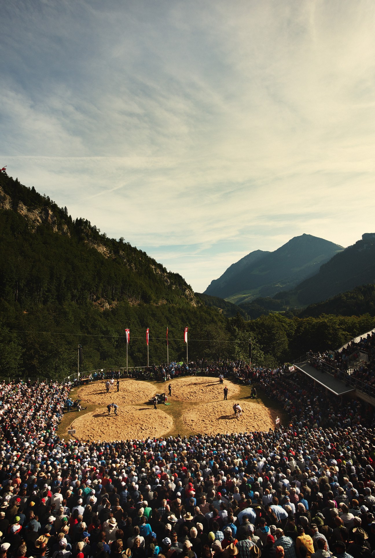 Large crowd of spectators surrounds multiple sawdust wrestling rings at Brünig's Schwingplatz, nestled in an Alpine valley.