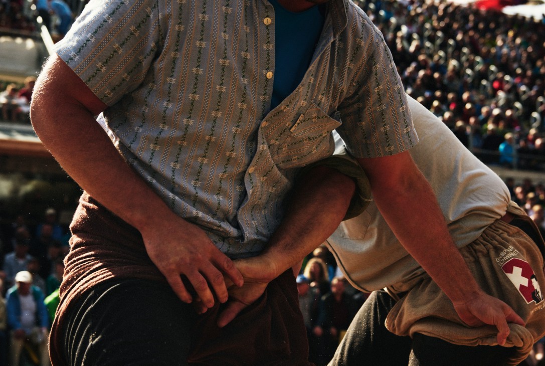 Two Swiss wrestlers grappling in traditional Schwingen wrestling attire with patterned shirts and burlap hose, spectators visible in background.