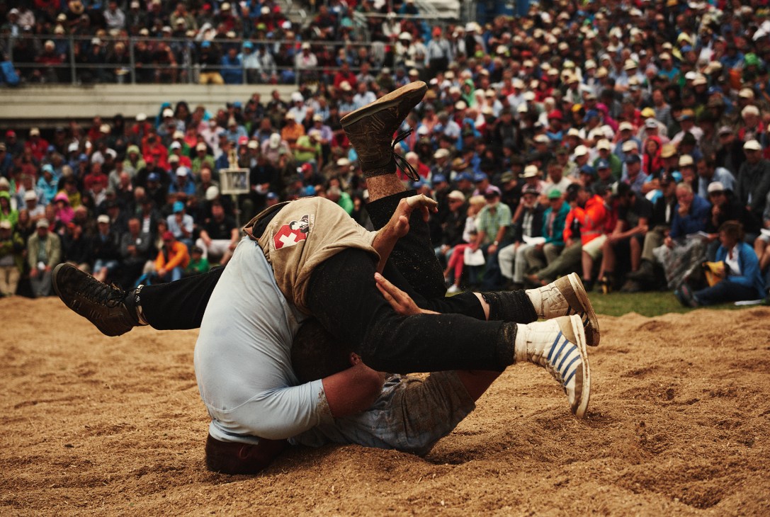 A Swiss wrestler lies on his back in sawdust during a traditional Schwingen match with spectators in the background.