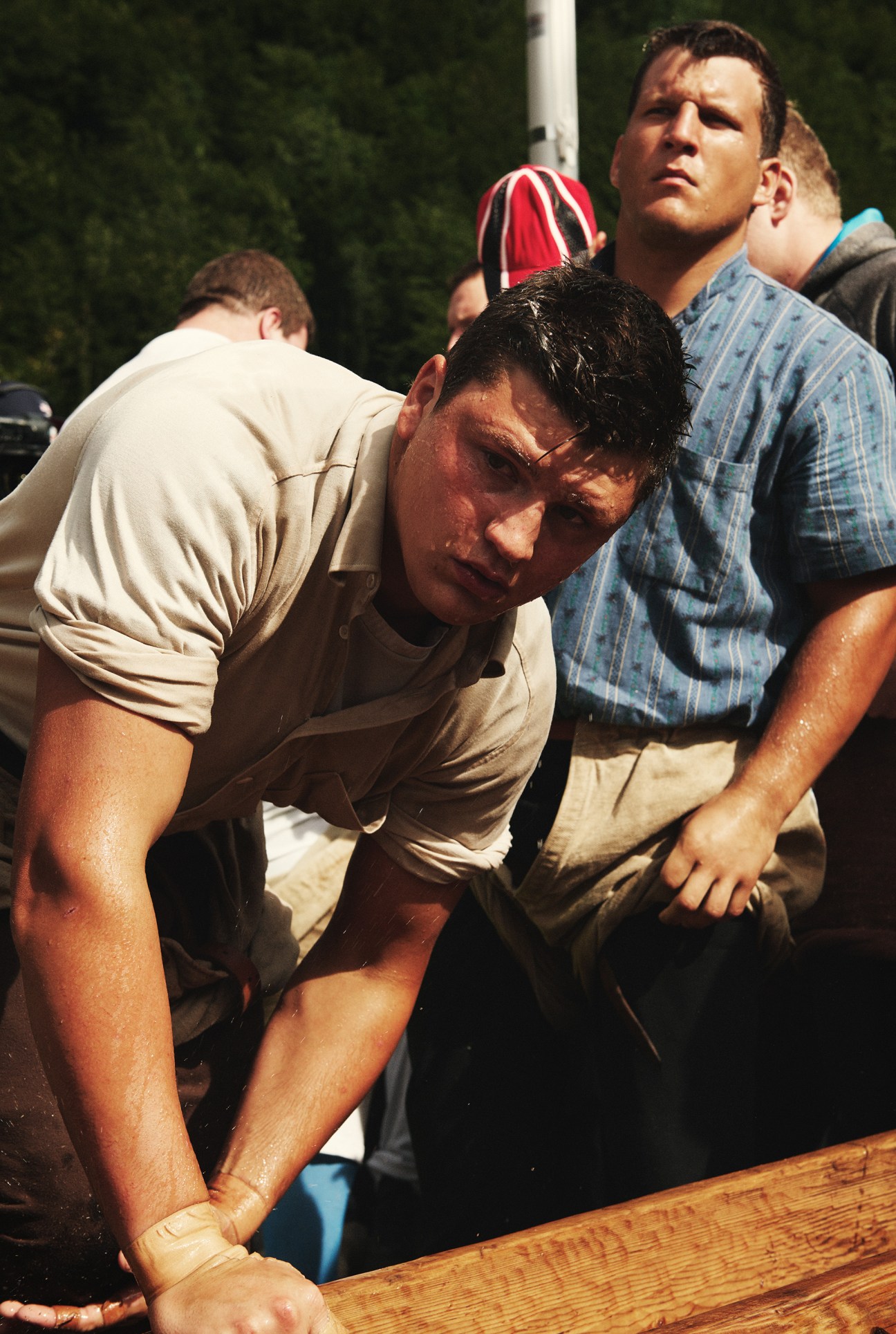 A muscular man in a beige shirt bends over a wooden trough while spectators watch during a Swiss wrestling event.