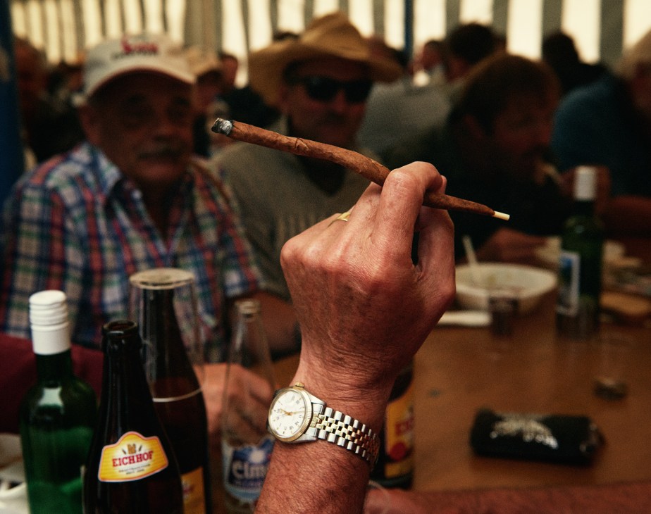 A weathered hand holding a cigar while watching traditional Swiss wrestling, with spectators and beer bottles visible in the background.
