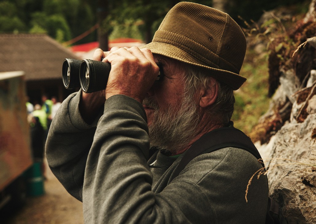 An older man wearing a hat and binoculars watches the Schwingen wrestling event from a vantage point at the Brünig Pass.
