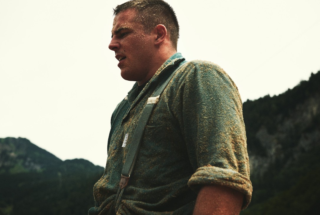 A Schwingen wrestler covered in sawdust after a match, standing against an Alpine mountain backdrop.