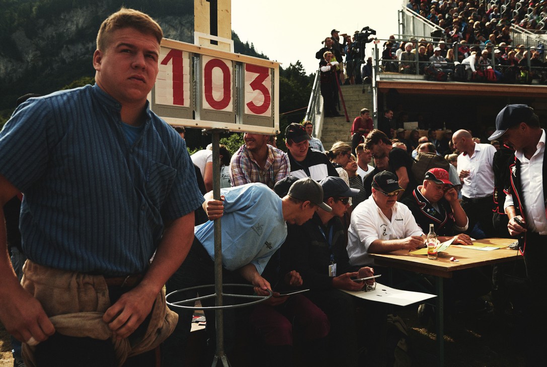 Man in blue shirt holds scoring board at Swiss wrestling event with judges and spectators.