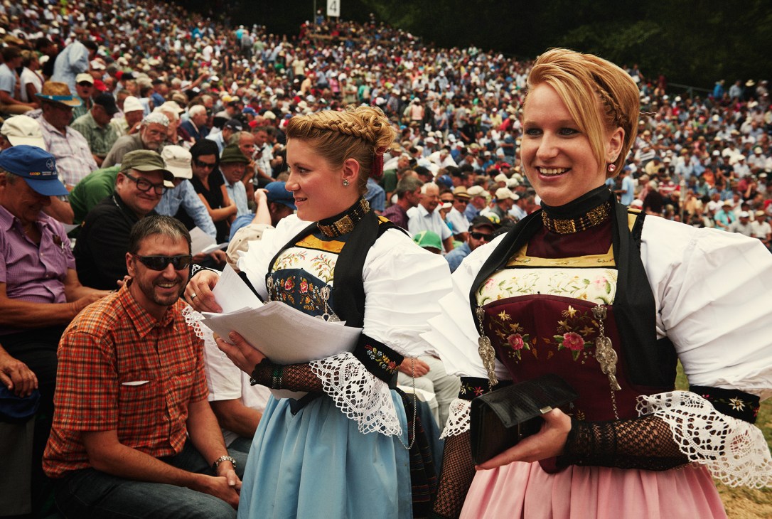 Two women in traditional Swiss dirndl dresses smile while holding a document at a crowded outdoor event.