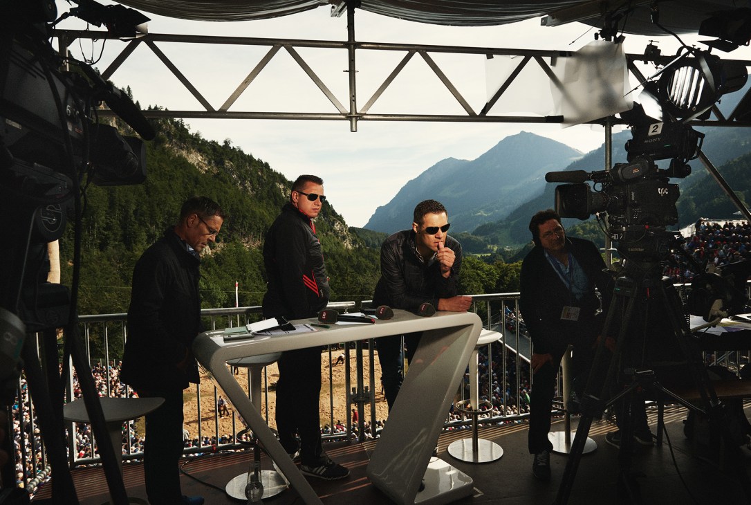 Four men in dark jackets sit at a broadcast table under scaffolding with Alpine mountains and crowds visible behind them.