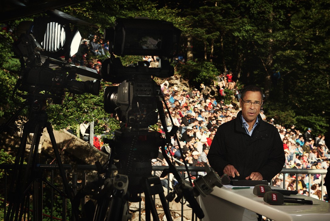 SRF television commentator at broadcasting desk with camera and crowd of spectators in background