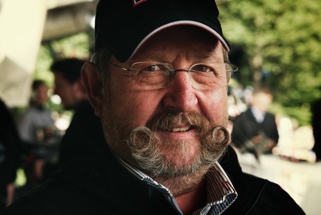 Edi Ritter wearing glasses and a black cap with a distinctive curled mustache, smiling at the camera outdoors.