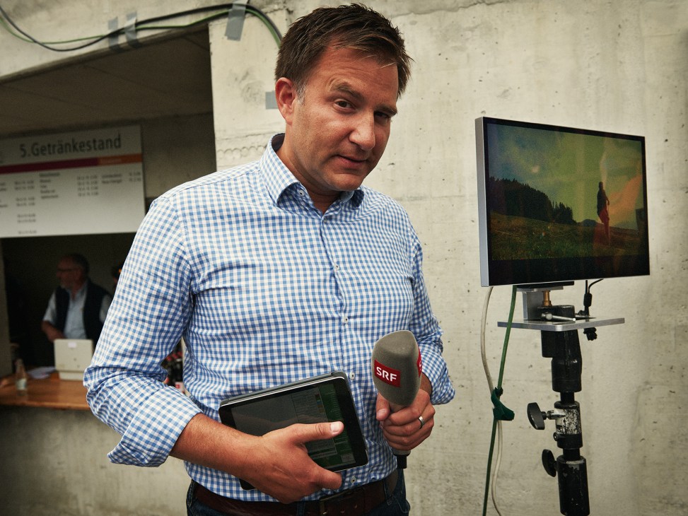 SRF commentator Sascha Ruefer holding a microphone and tablet in a broadcast booth.