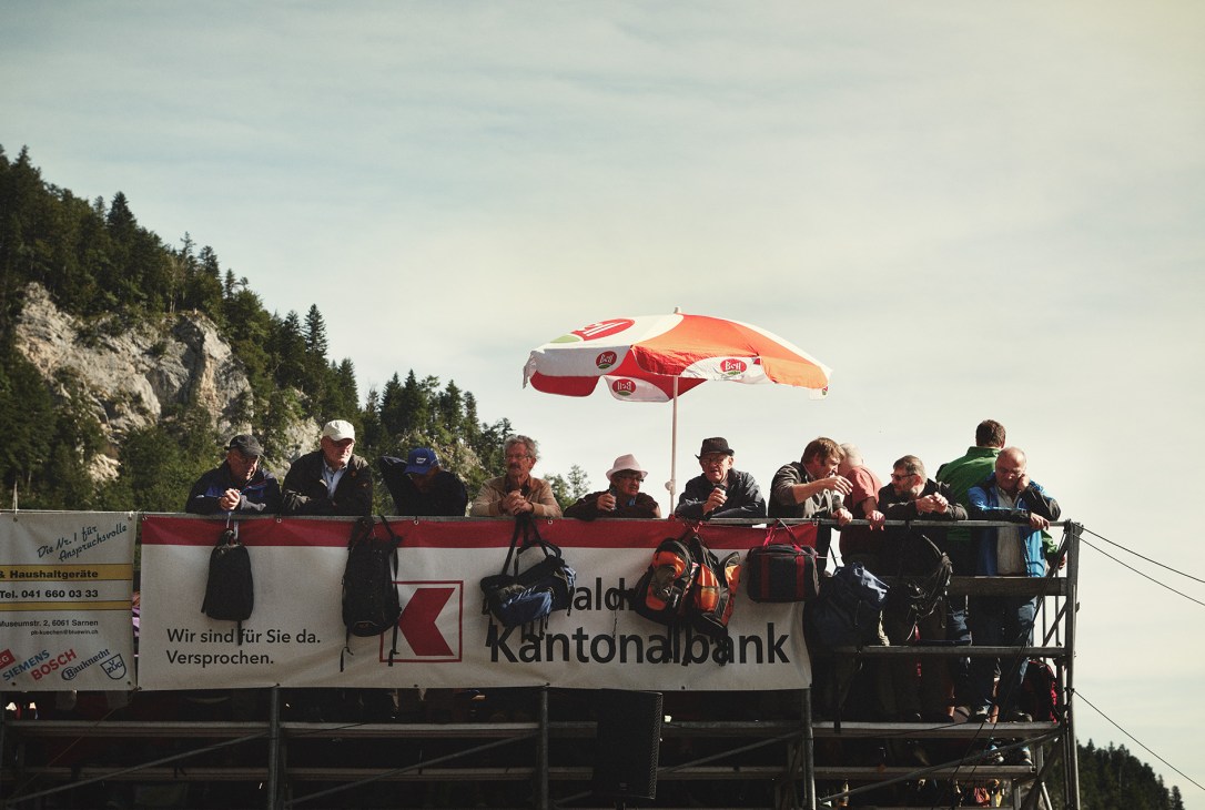 Spectators watch from a shaded viewing platform with a red and white umbrella at the Brünig Schwingen wrestling event in Switzerland.