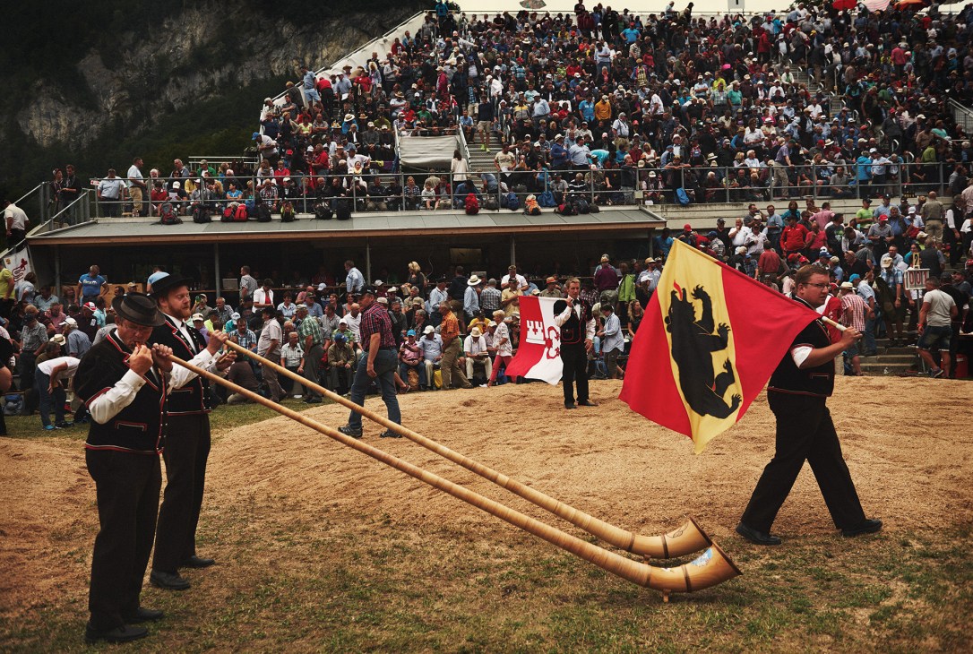 Two men in traditional Swiss attire play long alphorns while a flag bearer holds a red and yellow cantonal flag at the Schwingen wrestling event.