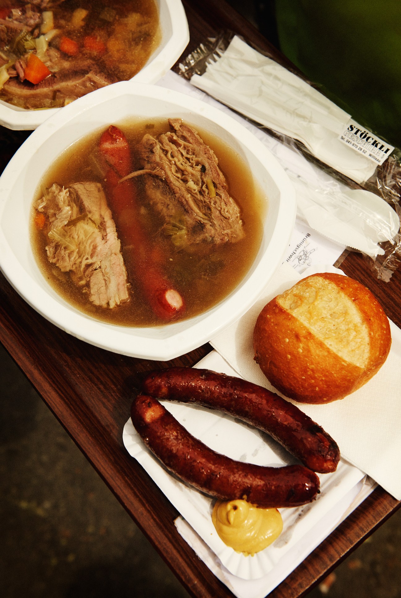 Overhead view of siedfleisch stew in white bowl with bratwurst sausages, bread roll, mustard and napkins on wooden surface.