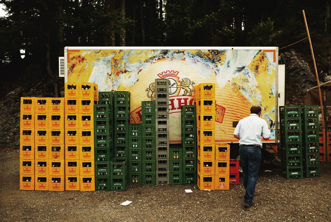 Man in white shirt standing before stacked beer crates with brewery signage and Alpine mural backdrop.