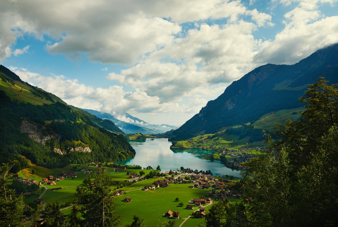Alpine valley landscape with Lungerersee lake surrounded by green meadows, forested mountains, and scattered chalets under cloudy sky.