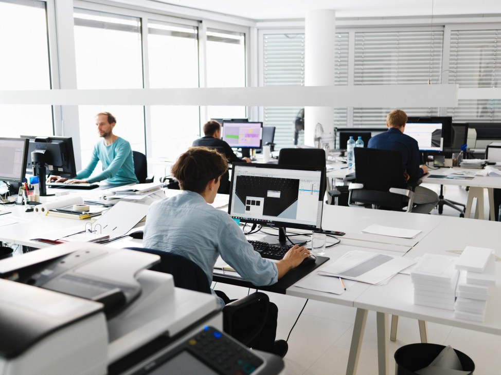 Multiple architects work at computer stations in a bright, modern DMAA office with large windows and white desks.